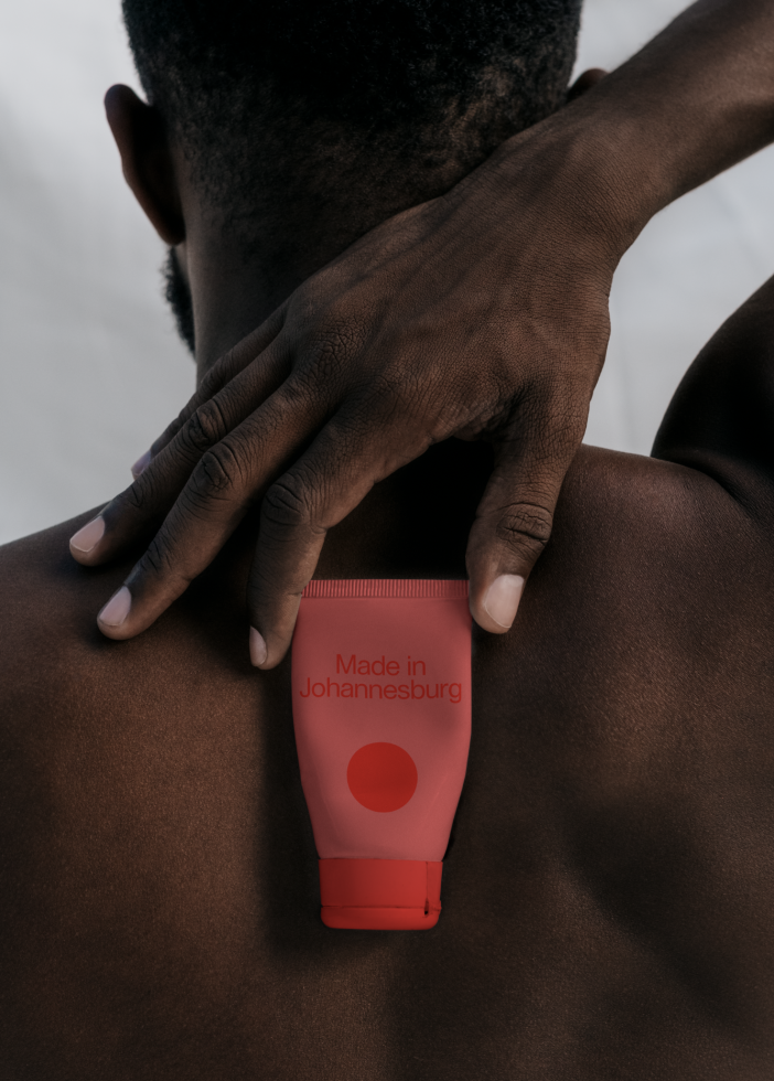 Person holding red product tube with text Made in Johannesburg on their shoulder, skin care packaging mockup for designers.